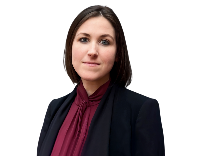 Professional woman with straight, shoulder-length dark brown hair, wearing a dark blazer over a maroon blouse with a tie-neck detail. She stands in front of a softly blurred light background, looking confidently into the camera with a composed expression.
