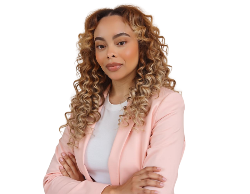 Confident woman with long, tightly curled ombré hair wearing a light pink blazer over a white top, standing with arms crossed. She poses against a softly blurred background with a poised and self-assured expression, blending professional style with a mode
