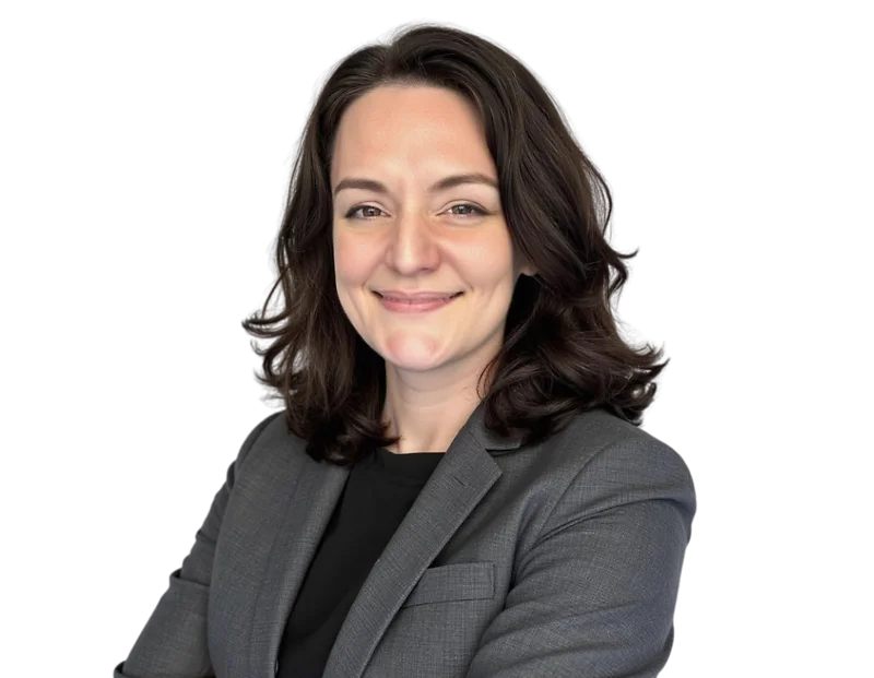 Smiling professional woman with shoulder-length dark wavy hair wearing a gray blazer over a black top, standing with arms crossed against a transparent background. Her expression is friendly and confident, projecting a composed and competent business pres