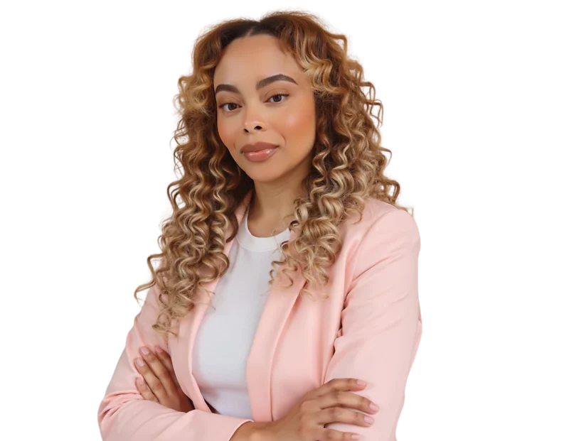 Confident woman with long, tightly curled ombré hair wearing a light pink blazer over a white top, standing with arms crossed. She poses against a softly blurred background with a poised and self-assured expression, blending professional style with a mode