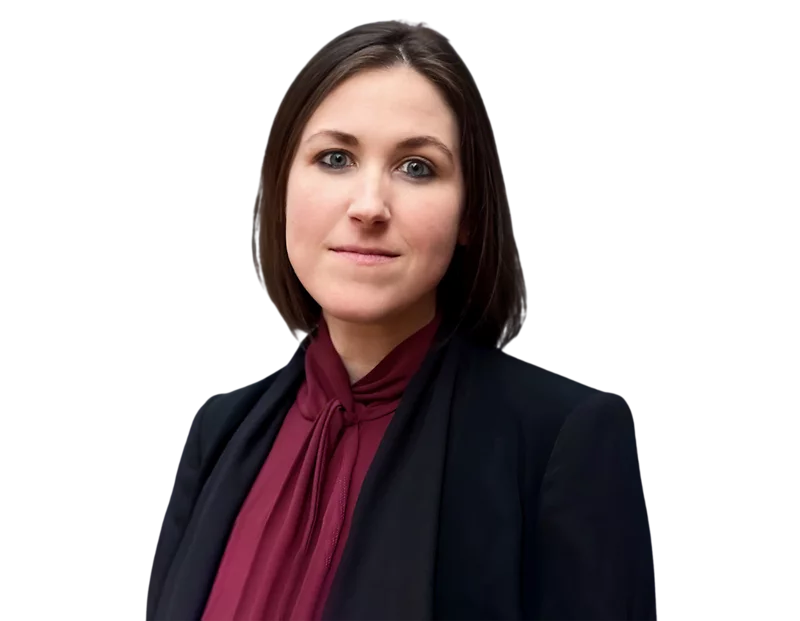 Professional woman with straight, shoulder-length dark brown hair, wearing a dark blazer over a maroon blouse with a tie-neck detail. She stands in front of a softly blurred light background, looking confidently into the camera with a composed expression.