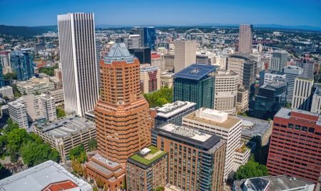 Aerial view of downtown Portland, Oregon, with a mix of modern skyscrapers and mid-rise buildings. The city is surrounded by greenery and distant hills under a clear blue sky.
