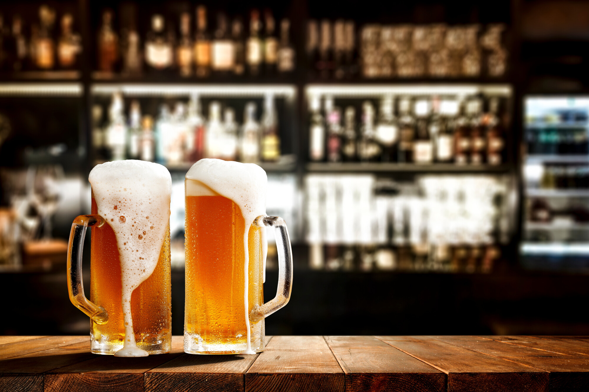 Two frosty glass mugs of golden beer with thick foam overflowing onto a wooden bar counter with a softly blurred bar and bottles in the background.