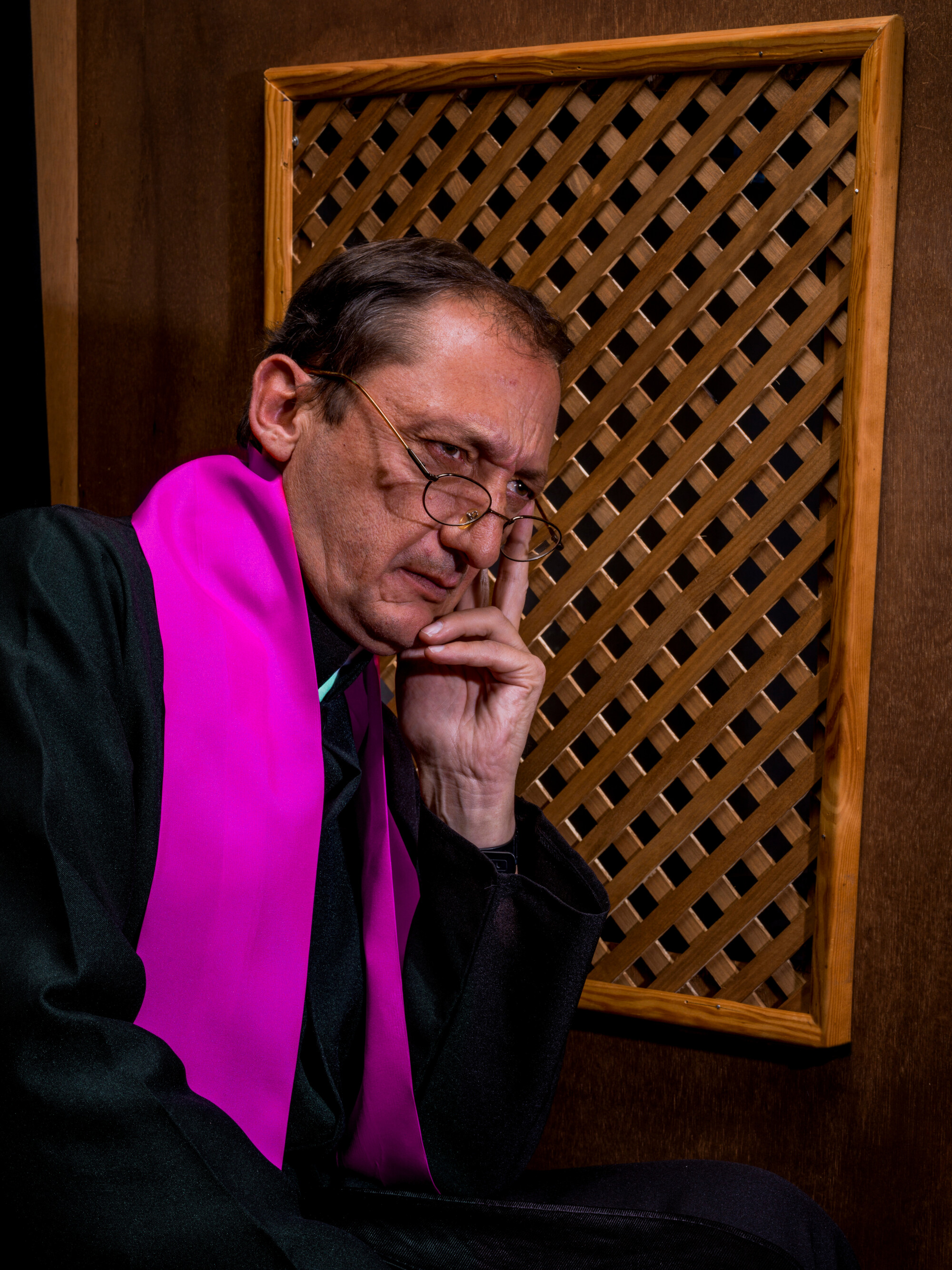 Man wearing a clerical robe and bright stole sits beside a wooden confessional screen with a serious and contemplative expression, resting his face on his hand.