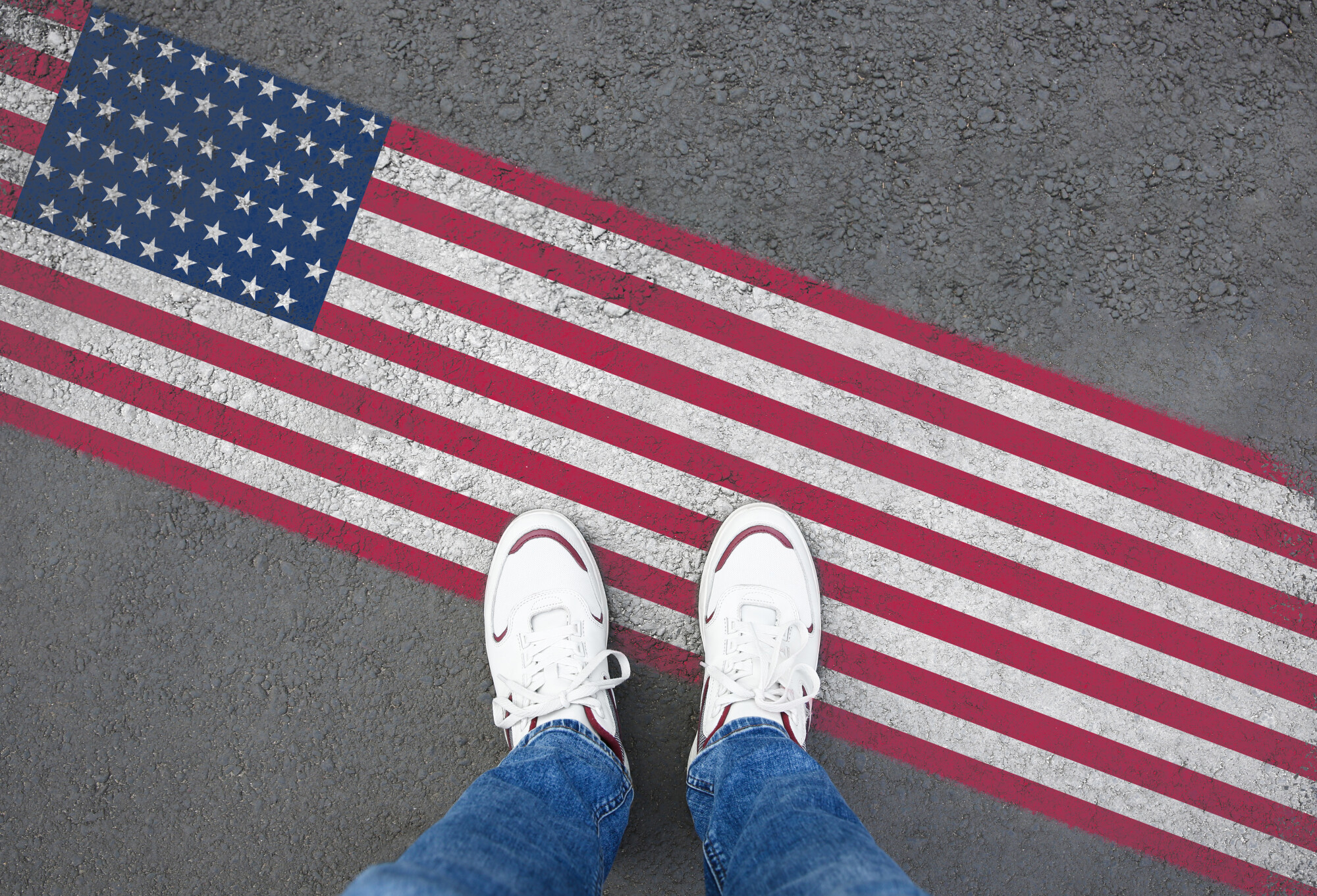 Person wearing white sneakers and jeans standing on asphalt next to a painted American flag stripe on the ground. The red white and blue flag design stretches diagonally across the pavement beneath their feet.