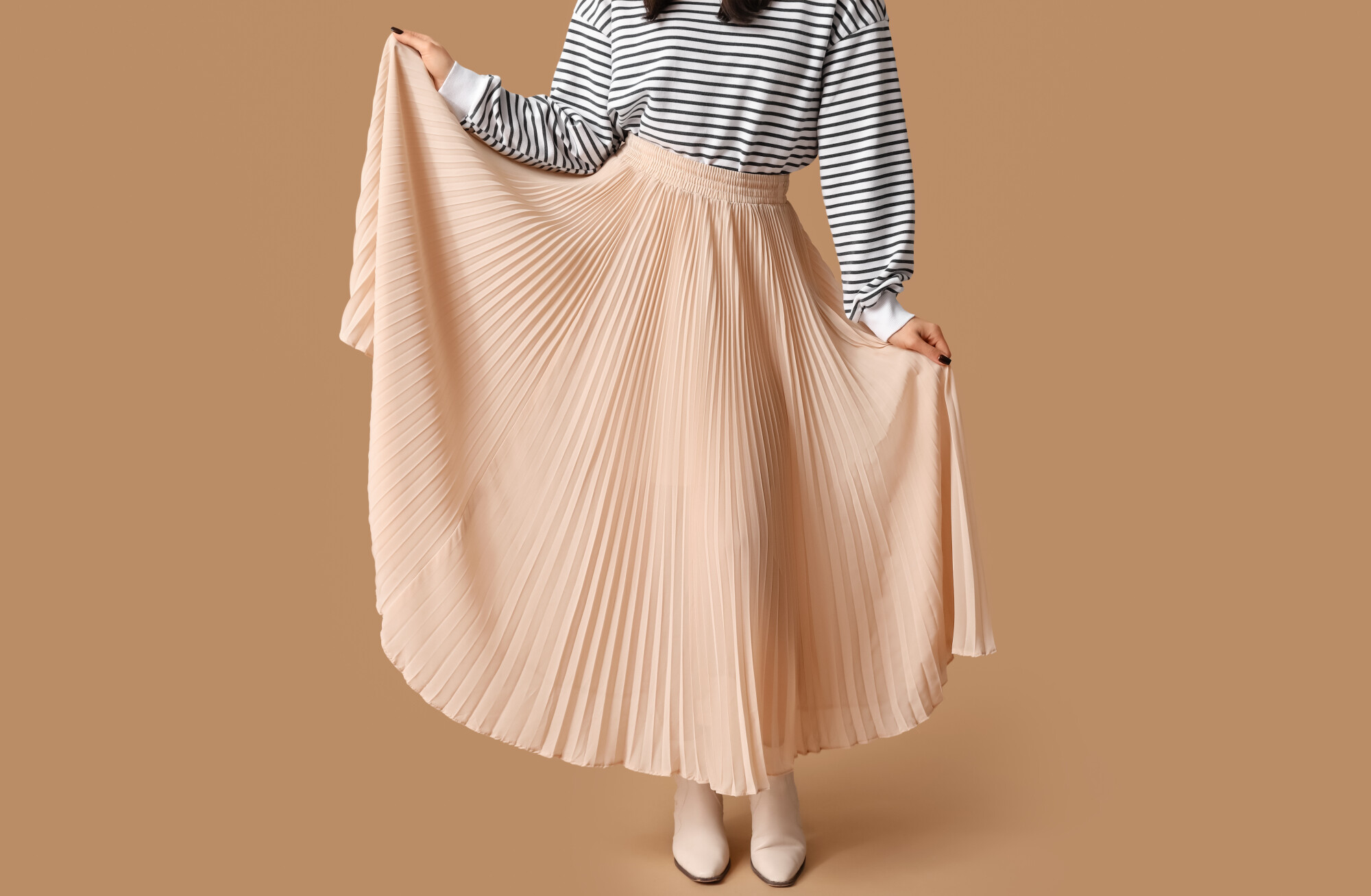 Woman wearing a black-and-white striped long-sleeve top paired with a flowing beige pleated midi skirt, holding out the sides to showcase the skirt's volume. She completes the outfit with white ankle boots, standing against a solid tan background.