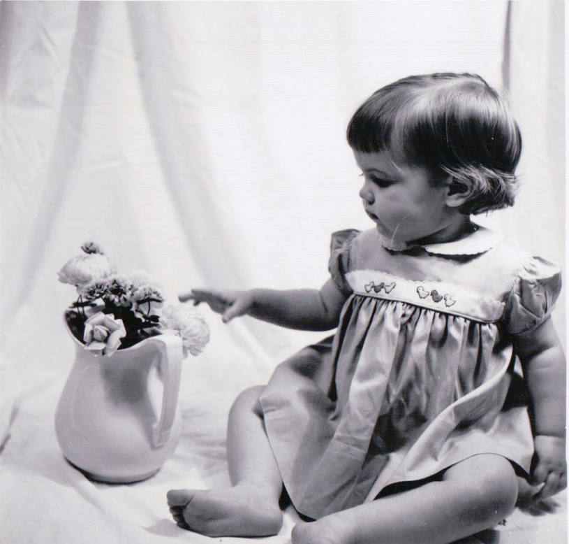 This is a black and white photograph of a young child sitting on a soft surface, reaching toward a ceramic pitcher filled with flowers. The child is wearing a dress with embroidered details and appears to be gazing curiously at the floral arrangement.        Ask ChatGPT