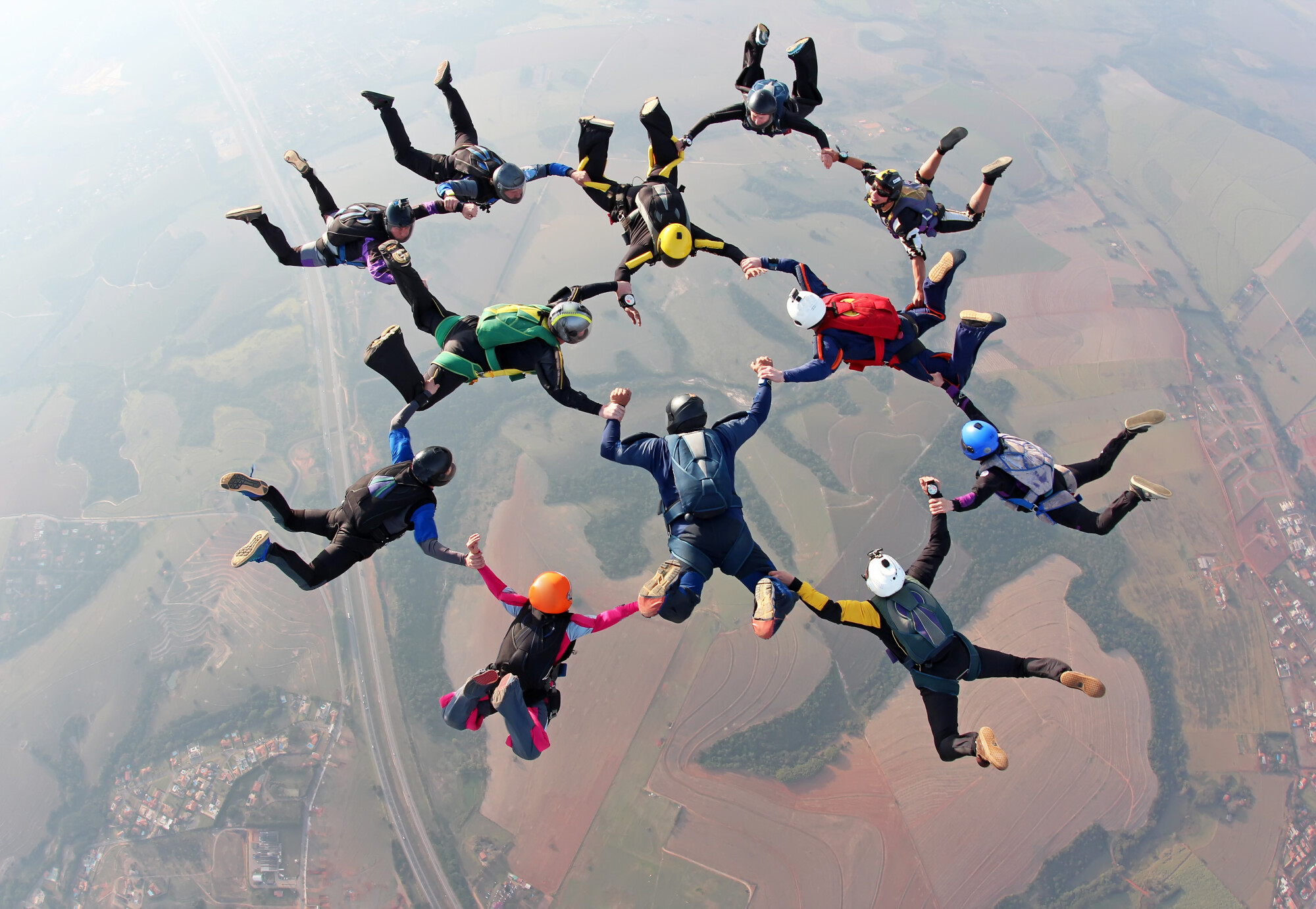 A group of skydivers in colorful jumpsuits and helmets form a large circle mid-air by holding hands during freefall. The coordinated formation showcases teamwork and precision against a backdrop of fields and roads far below.