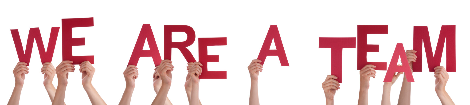 Multiple hands hold up large red letters spelling out "WE ARE A TEAM" against a white background. The coordinated effort visually emphasizes unity, collaboration, and teamwork.
