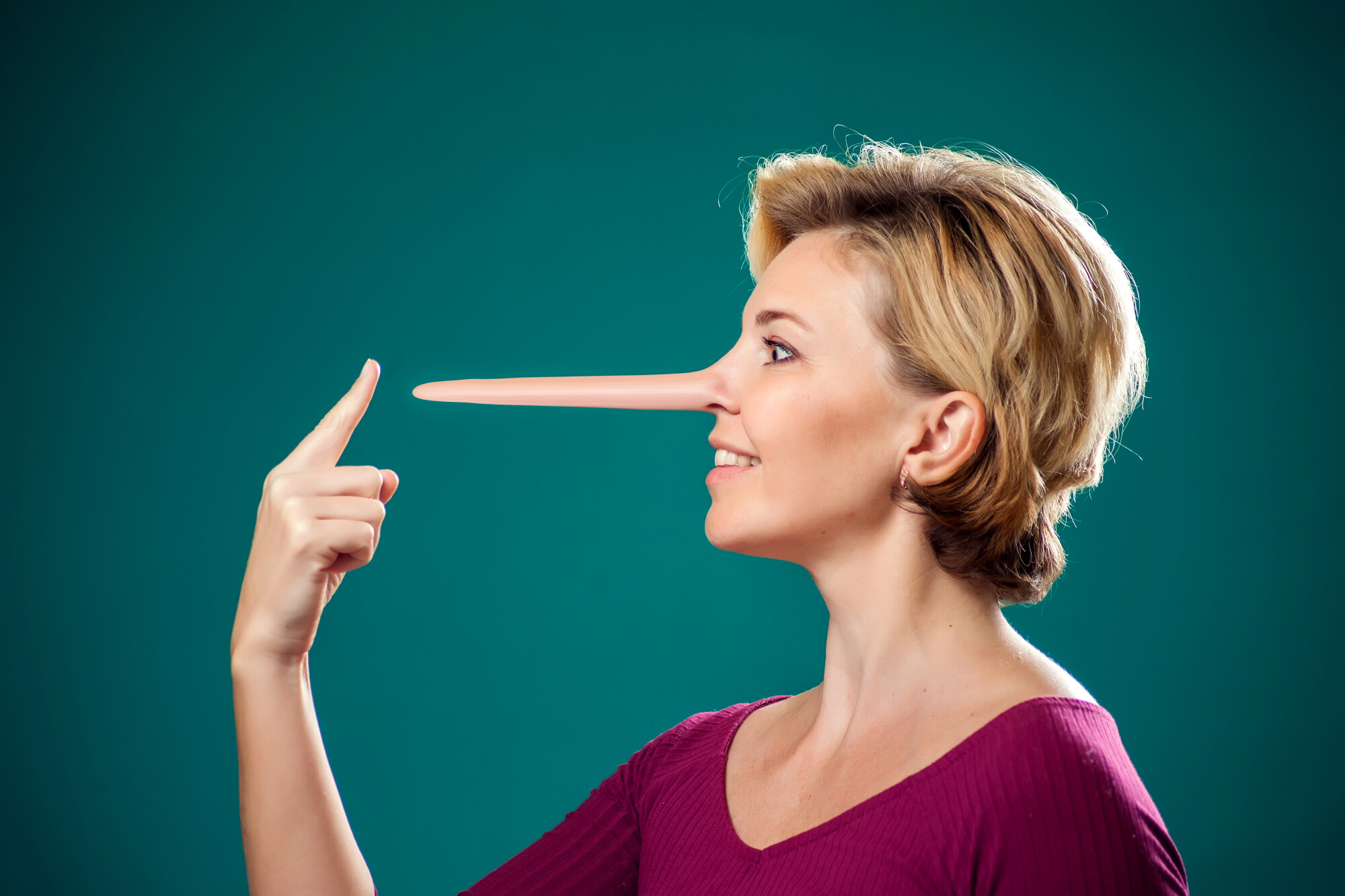 A woman with short blonde hair smiles while pointing at her unnaturally long nose, a visual reference to Pinocchio and the concept of lying. The green background and her purple shirt create a vivid contrast that emphasizes the humorous and exaggerated expression.