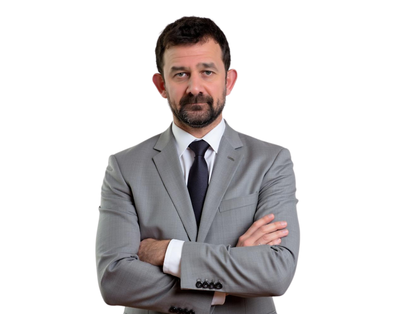 Serious-looking man with short dark hair and beard wearing a light gray suit and dark tie, standing with arms crossed against a transparent background. His formal attire and posture suggest a professional or executive portrait.