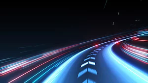 Futuristic illustration of a curving road with glowing blue and red light trails, suggesting high-speed motion through a dark environment. The dynamic perspective conveys themes of technology, innovation, and rapid progress.