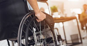 A disabled man is sitting in a wheelchair. He holds his hands on the wheel. Nearby are his colleagues