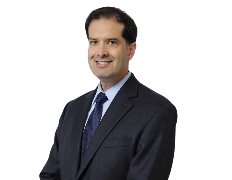 Colin Thakkar in a dark suit and blue tie stands smiling against a modern blue and white geometric background.