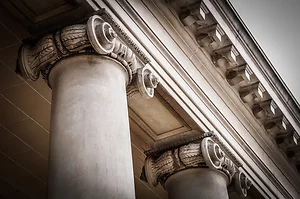 Close-up view of classical architectural columns with ornate Ionic capitals, featuring scroll-like volutes and carved detailing. The weathered stone and perspective highlight the elegance and strength of historic building design.