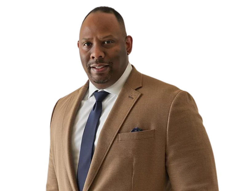 Darrick McDuffie in a tan suit jacket, white shirt, and navy tie, standing against a light blue and white background.