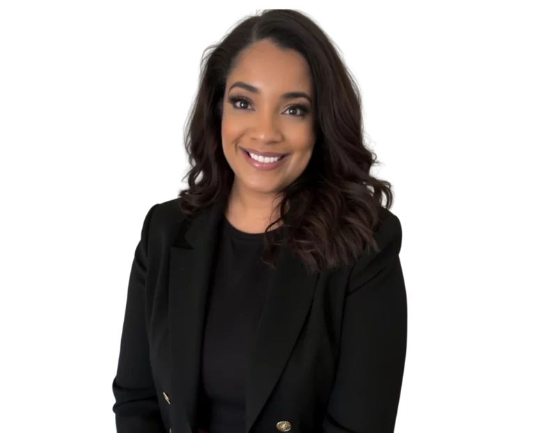 Professional headshot of Taren Greenidge with shoulder-length wavy hair wearing a black blazer and black top, posed against a plain background. She appears confident and professional, with a warm expression and natural makeup.