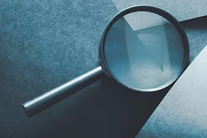 Close-up of a magnifying glass resting on textured blue paper with layered geometric shapes. The lens slightly distorts and enlarges the paper’s surface, creating a subtle play of light and shadow.