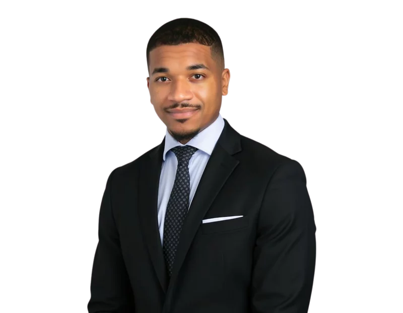 Professional headshot of a young man in a black suit with a white shirt and dark patterned tie, standing against a white background and smiling slightly. His look is polished and business-ready, with neatly groomed hair and a confident posture.