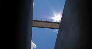 Looking up between two tall dark concrete walls toward a narrow pedestrian bridge crossing overhead against a bright blue sky. The sun shines beside the bridge creating a lens flare, while small white clouds drift through the open space above.