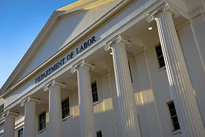 The image shows the facade of the U.S. Department of Labor building, featuring tall white columns in a classical architectural style. Sunlight casts long shadows across the structure, highlighting the fluted detailing and the bold black lettering of the b