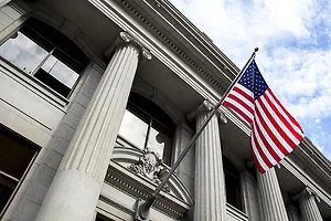 Low-angle view of a neoclassical stone building with tall fluted columns and an American flag waving in the foreground. The architectural details and patriotic symbol convey themes of government, law, or public institutions.