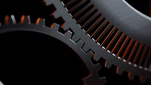 Close-up view of two interlocking metallic gears with sharp, polished teeth. The edges are highlighted by a warm orange light, contrasting with the dark, shadowed background.