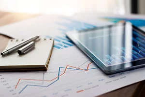 The image shows a close-up of a desk with a tablet, a pen, and a notepad placed on printed charts featuring line and bar graphs. The graphs display fluctuating data trends in red, blue, and gray, suggesting a focus on financial or business analysis.