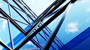 Upward view of a modern glass and steel building with angular beams reflecting the blue sky and scattered clouds. The structure's geometric lines create a dynamic, futuristic perspective.