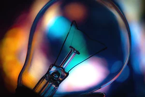 Close-up photograph of a clear light bulb against a blurred background of vivid blue, orange, and purple bokeh lights. The bulb’s delicate filament and glass structure are sharply in focus, contrasting with the soft, colorful glow behind it.